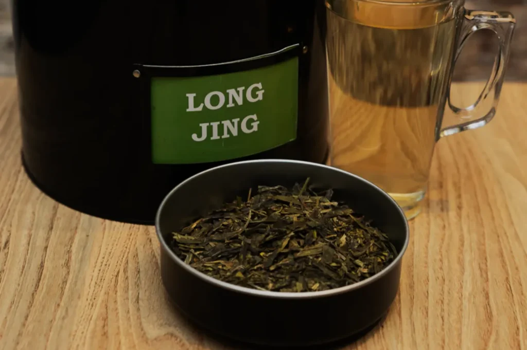 A tin labeled "Long Jing" is next to a bowl of green tea leaves and a clear glass mug filled with steeped tea, set on a wooden surface.