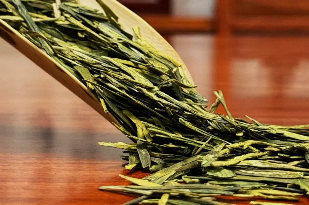 Loose green tea leaves spill from a wooden scoop onto a smooth table. The leaves are vibrant, creating a fresh and aromatic atmosphere.