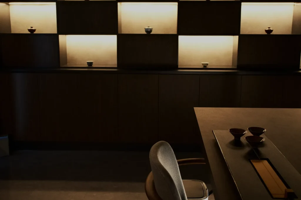 Modern interior with a minimalist design. Wooden shelves with soft lighting display ceramic bowls. A gray chair and table create a serene, elegant ambiance.