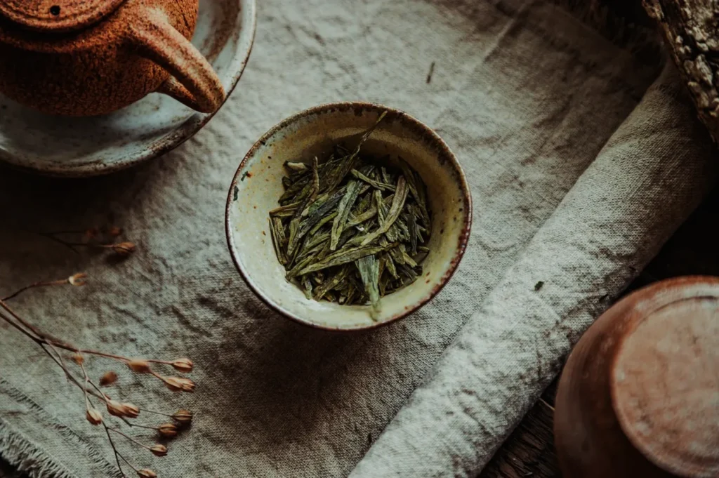 A rustic setting with dried green tea leaves in a ceramic bowl, a textured beige cloth underneath, and a matching teapot nearby, evoking a cozy, natural feel.