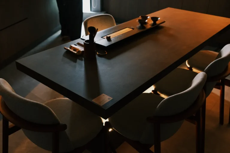 A dimly lit dining table with a dark surface and surrounding chairs, featuring place settings and minimal decor.