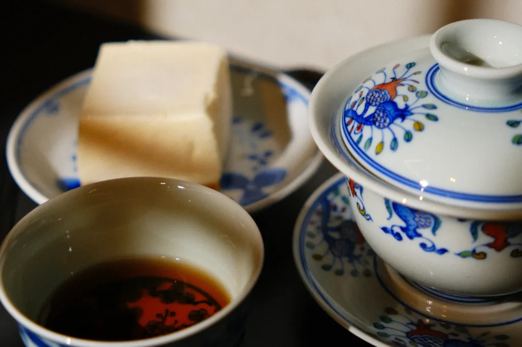 Porcelain teacup and teapot with blue floral designs, a cup of tea, and a block of tofu on a matching plate, creating a serene, traditional scene.