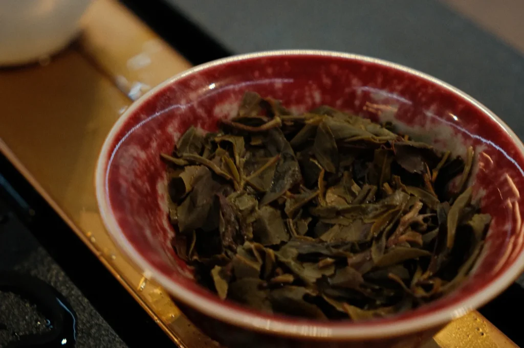 A red bowl filled with green tea leaves sits on a wooden tray. The leaves appear wet and slightly unfurled, suggesting they have been steeped.