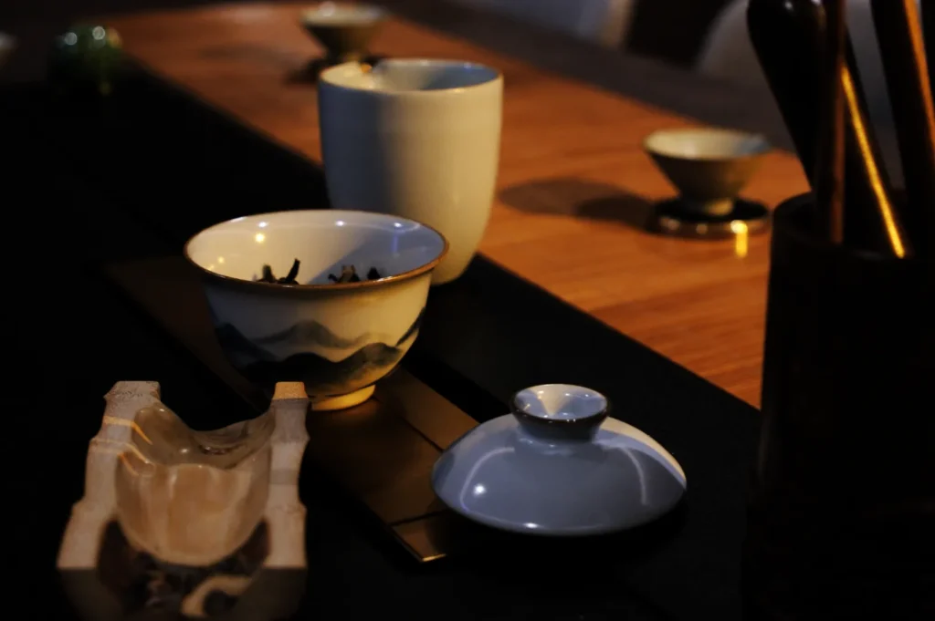 A tea set arranged on a wooden table, featuring a small tea cup with tea leaves inside, two more tea cups at the back, and a teapot, with a warm lighting ambiance.