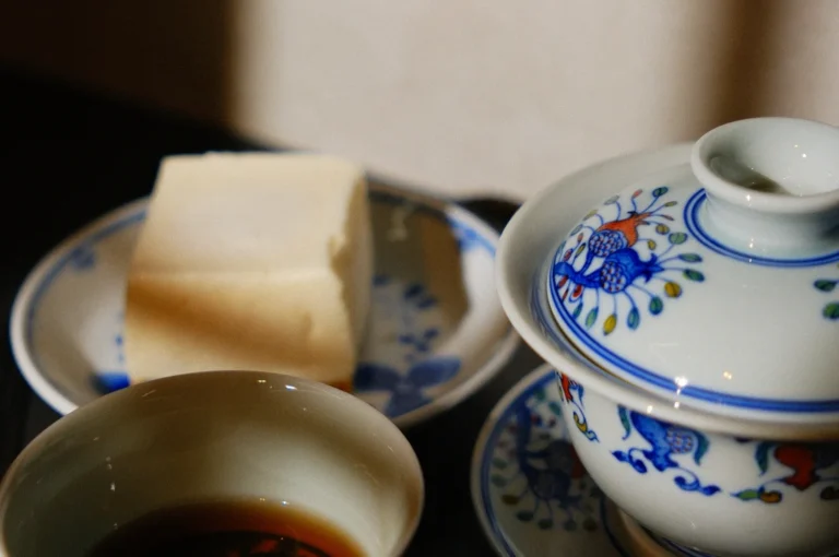 A piece of cheesecake on a decorative plate next to a small teacup and a lidded teapot, all featuring floral designs.