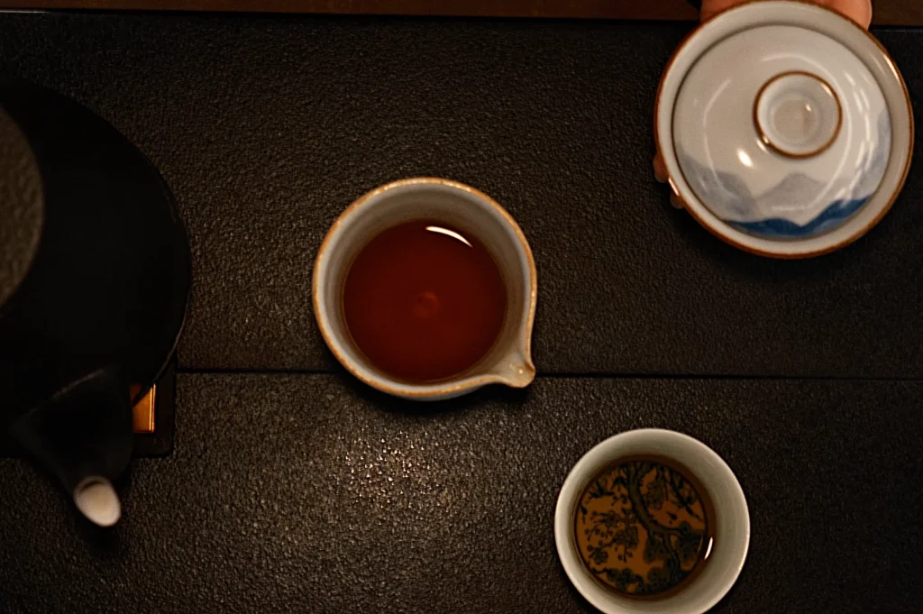 A teapot and two cups of tea on a dark surface. One cup has clear tea while the other contains tea with visible design from the bottom of the tea cup.