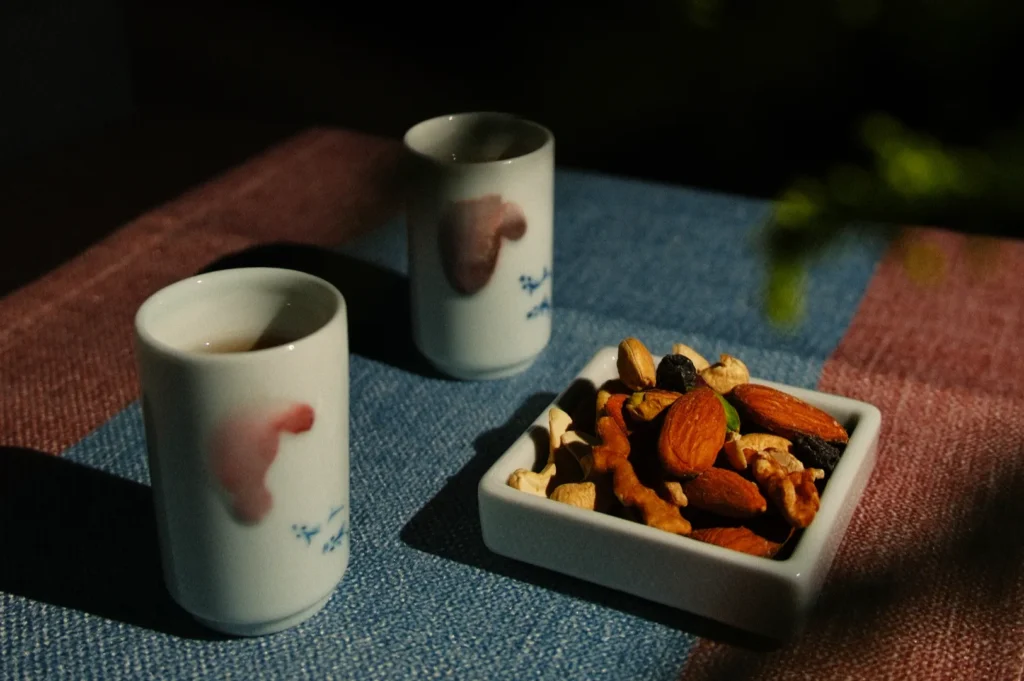 Two ceramic cups with watercolor designs and a rectangular dish of mixed nuts are placed on a textured tablecloth, lit by soft, diffused light.