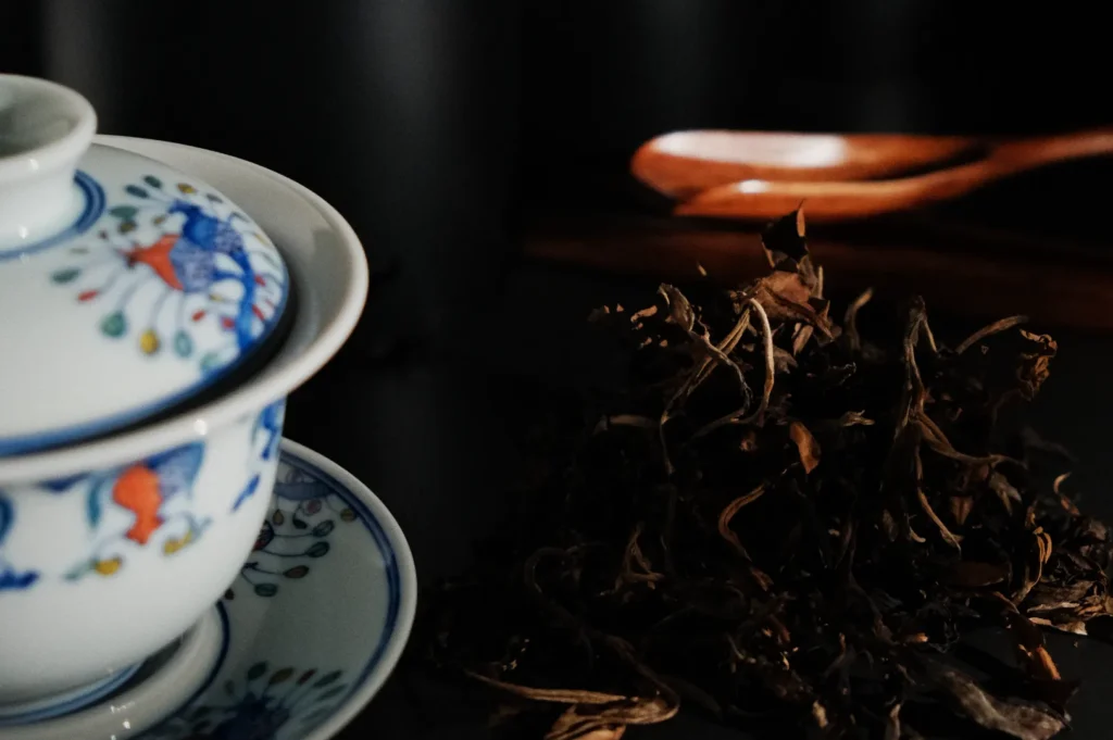 A blue and white ceramic teacup and saucer are placed near a mound of dried tea leaves, with a wooden spoon partially visible in the background. The setting features a dark surface, accentuating the colors of the teacup and the texture of the tea leaves.