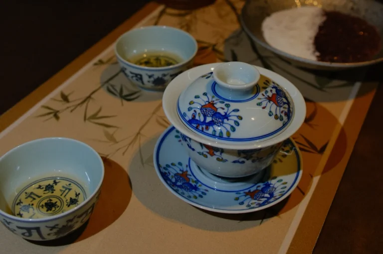Porcelain tea set with floral designs on a bamboo-patterned mat. Two cups contain tea, while a covered bowl and a dish with red bean paste are nearby.