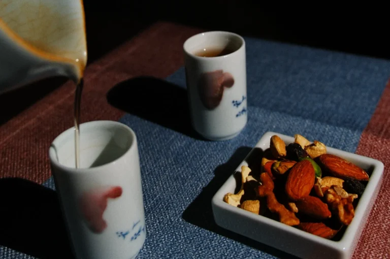 Tea is being poured into a small white cup with a subtle red design, beside another filled cup and a dish of mixed nuts on a textured mat. Cozy and warm atmosphere.