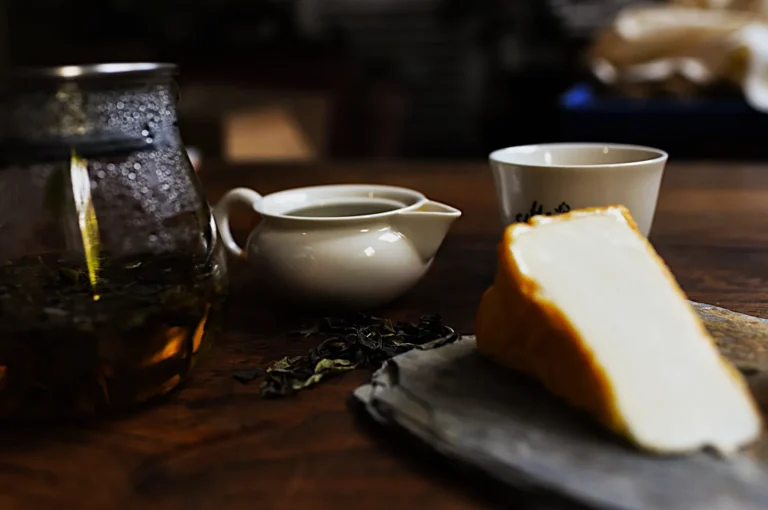 A cozy scene with a glass teapot of brewed tea, white porcelain creamer and cup, dry tea leaves, and a wedge of cheese on slate, set on a wooden table.
