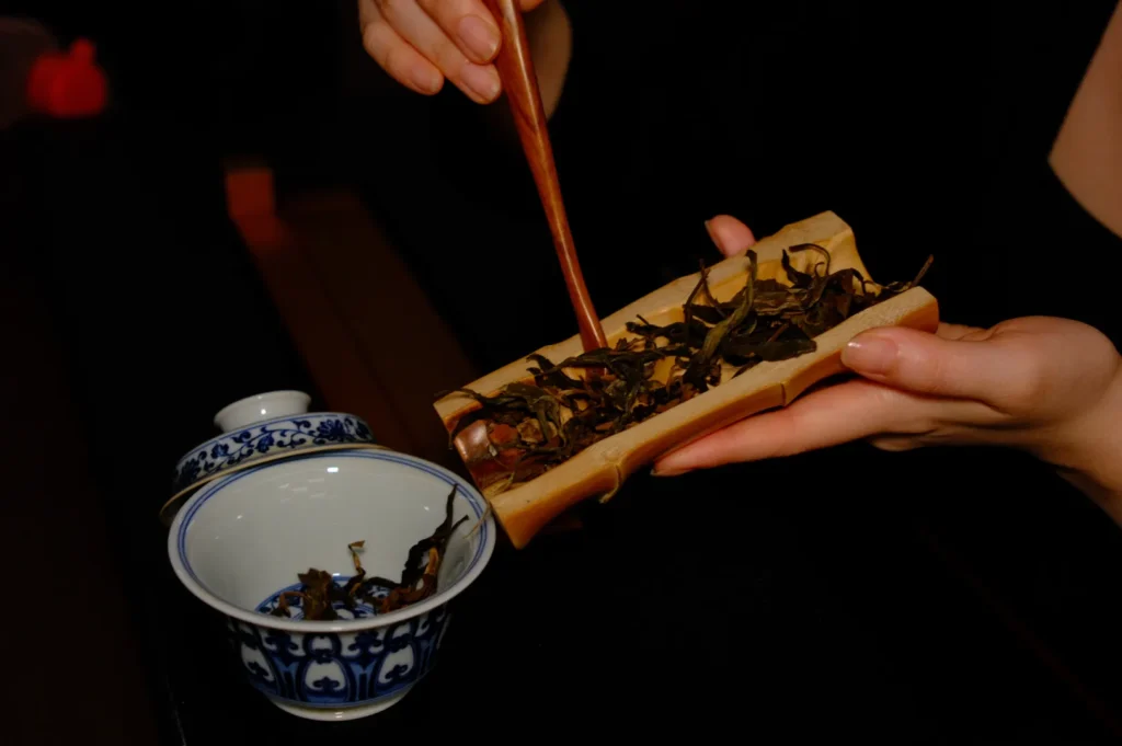 Hands delicately place tea leaves into a traditional blue and white cup using wooden tweezers, set against an elegant, dimly-lit background.