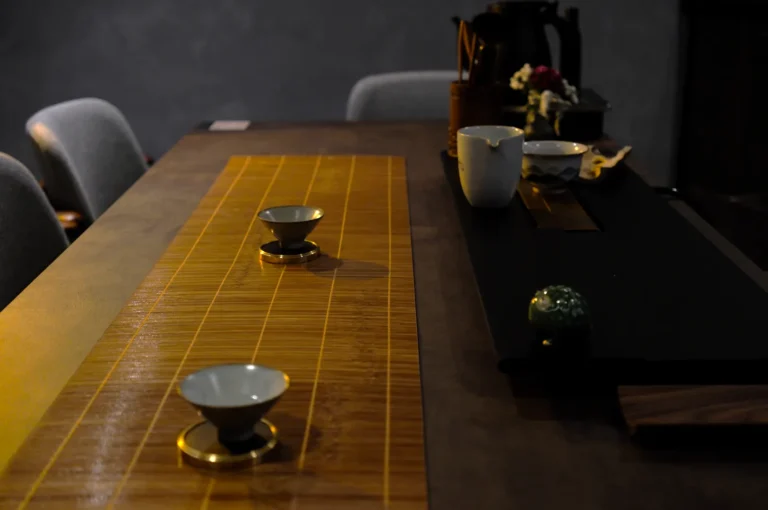 Dimly lit table set for tea, featuring two small cups on a bamboo mat. Chairs and a teapot create a calm, intimate atmosphere.