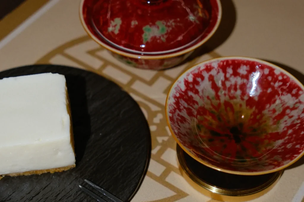 A square slice of cheesecake on a black plate beside two red, patterned ceramic bowls. The setting is elegant and artfully arranged.
