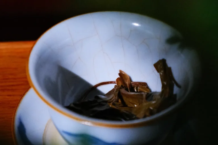 A close-up of a ceramic tea cup containing loose tea leaves, with soft shadows and natural light.