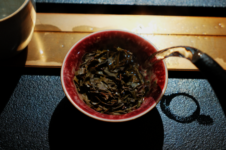 Pouring hot water into a red bowl filled with tea leaves, casting a warm, cozy atmosphere on a textured black surface beside a light wooden board.
