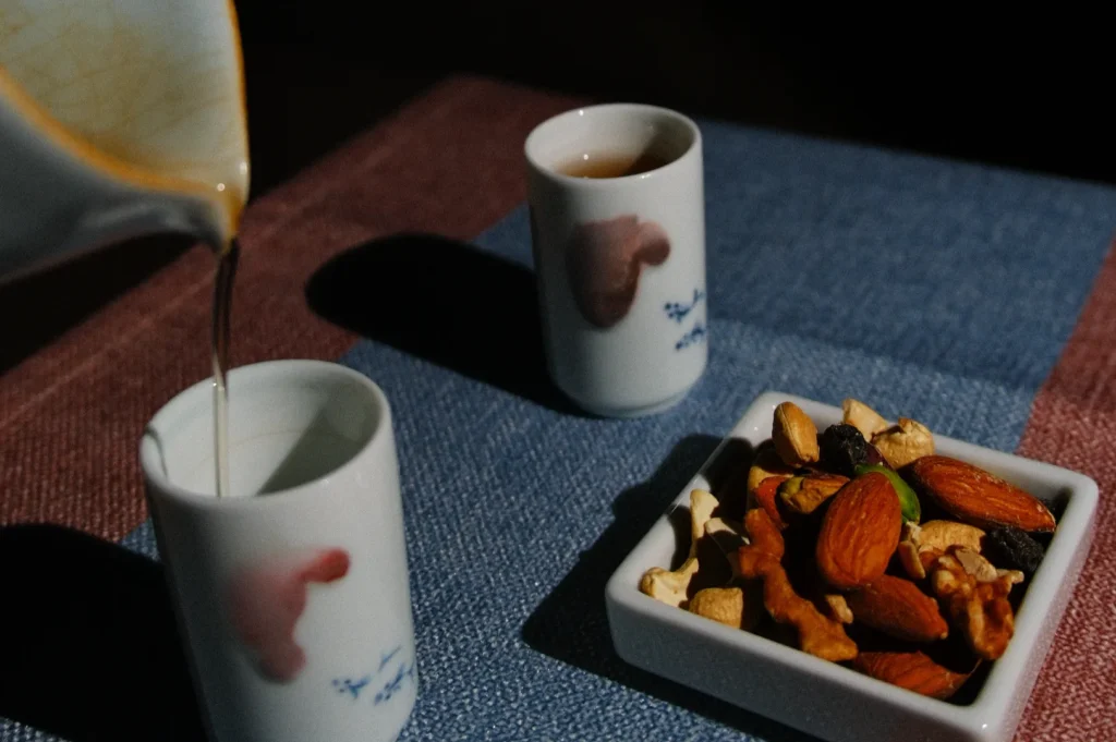 Ceramic pitcher pouring tea into a cup beside another filled cup. A bowl of mixed nuts and dried fruits rests on blue and pink fabric, creating a cozy vibe.