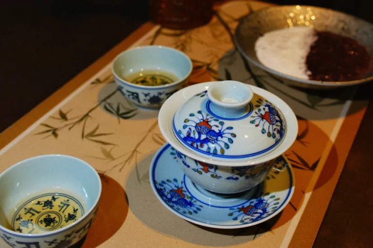 A traditional tea set with a decorative teapot and two tea cups, placed on a patterned tray surrounded by ingredients for tea preparation.