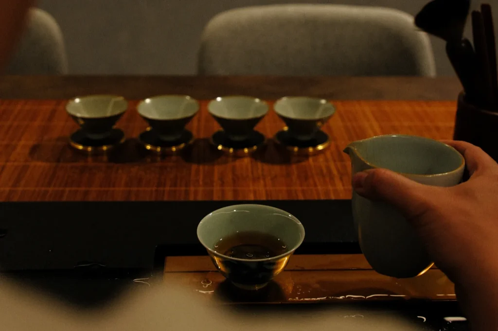 A person holding a teapot pouring tea into a small cup, with several empty cups arranged in the background.