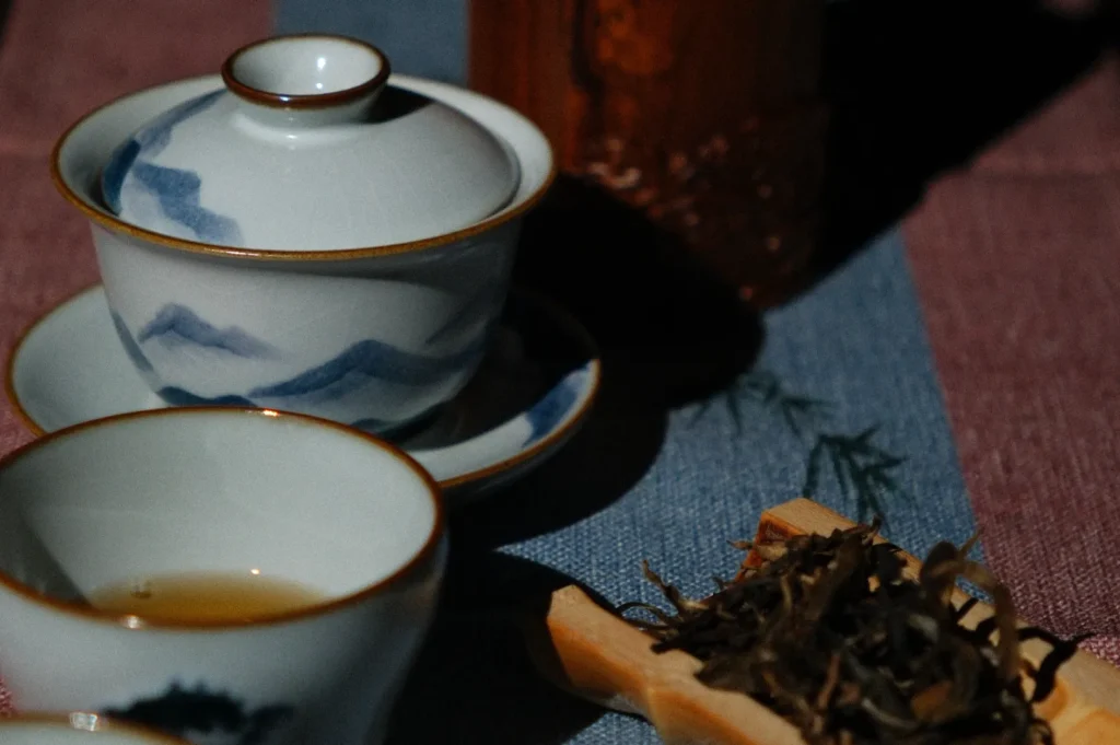 A close-up of traditional tea cups on a textured table setting, with loose tea leaves displayed in a wooden tray nearby.