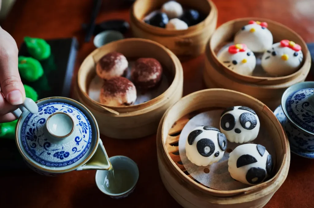 A hand pours tea from a blue-patterned teapot into a cup. Several bamboo steamers hold panda-shaped and decorated buns, creating a playful, cozy atmosphere.