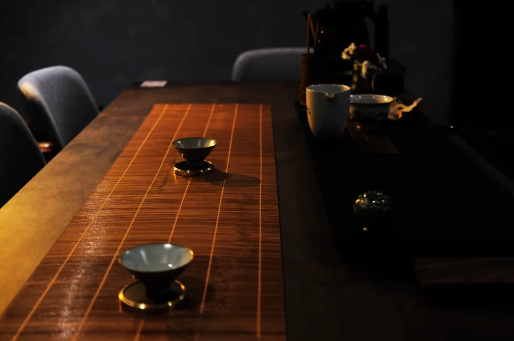 A dimly lit table setting with a wooden runner, featuring two small tea cups and a few other tea accessories in the background.
