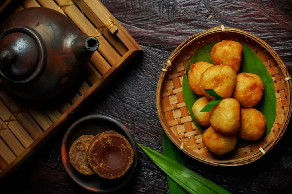 A woven bamboo basket filled with golden-brown fried dumplings, placed on a green leaf, accompanied by a small bowl of round cookies and a traditional teapot on a bamboo mat.