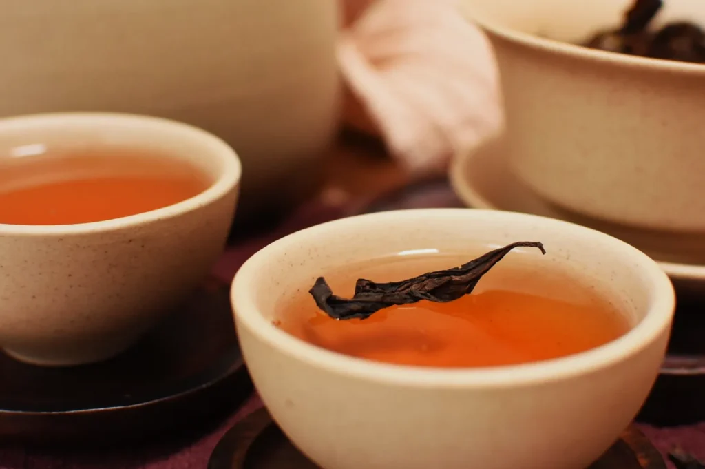 A close-up of two cups of tea, one with a single tea leaf floating on the surface, with additional tea pots in the background.