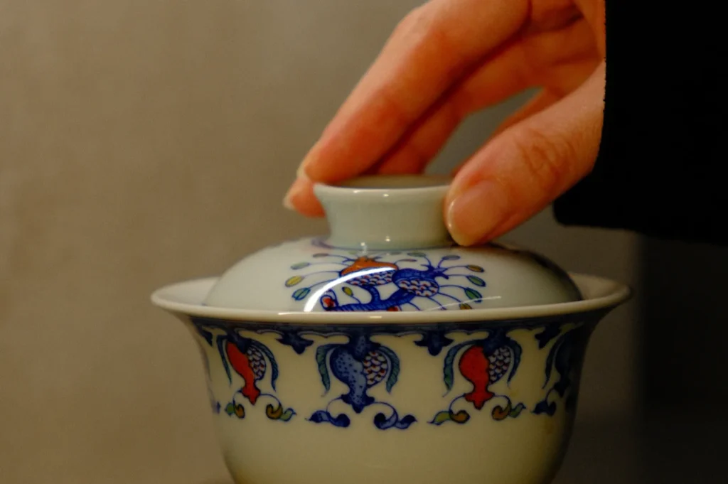 A hand wearing black long sleeves reaching for a white ceramic tea cup with a lid, featuring a decorative and colorful pattern.
