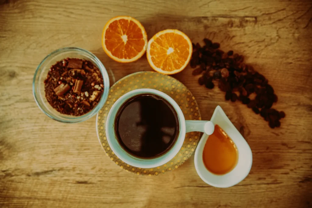 A cup of black coffee on a saucer, surrounded by a bowl of dried ingredients, two orange halves, honey in a dish, and scattered raisins on wood.