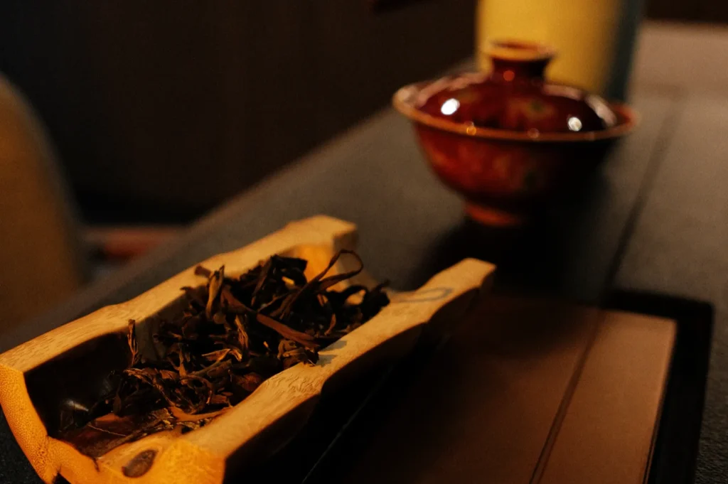 A wooden tray with dried tea leaves on it, with a red ceramic tea cup in the background.