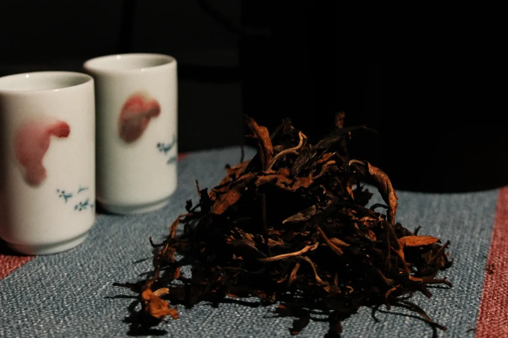 A pile of dried tea leaves is placed on a textured blue and red table cloth. In the background, two small white teacups with pink patterns are visible. A black kettle is partially visible behind the cups.