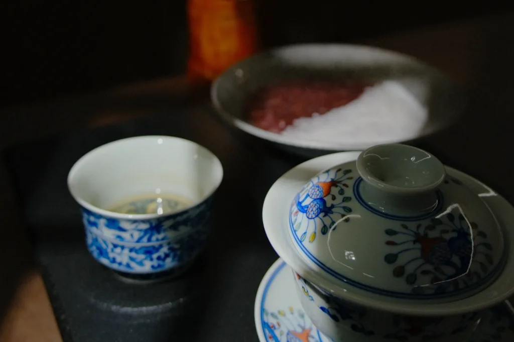 Ceramic tea set with floral designs on a dark surface; a small cup filled with tea and a covered bowl. Soft lighting adds a serene, inviting ambiance.