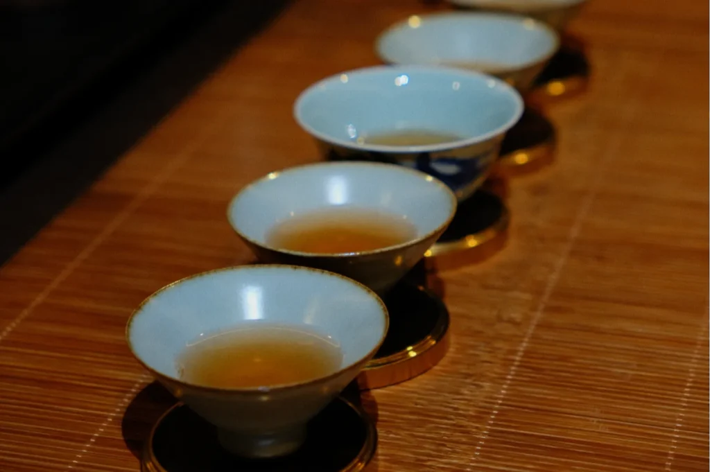 A row of five ceramic tea cups on a bamboo mat, each filled with pale tea. The setup conveys a sense of calm and traditional elegance.