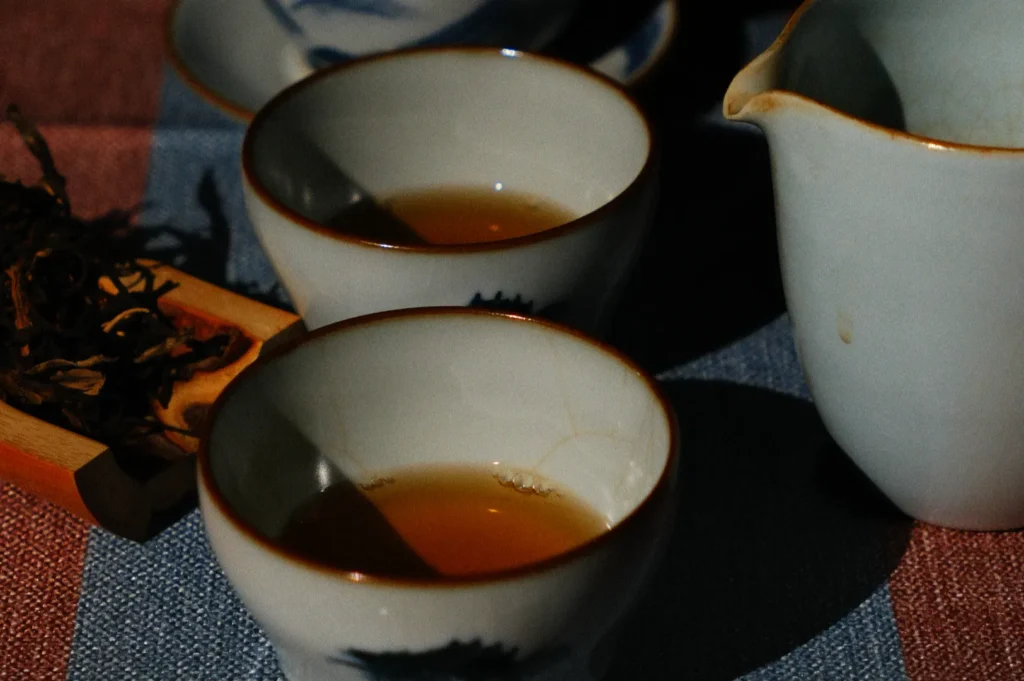 Two ceramic cups filled with amber tea sit next to a small white pitcher and a wooden dish of dried tea leaves on a striped cloth. The scene is cozy and warm.