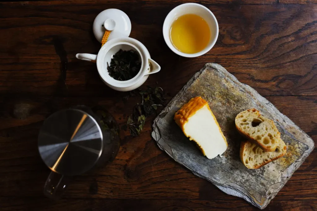 A rustic wooden table with a slate board holding a slice of cheese and two pieces of bread. Nearby, a teapot, tea leaves in a strainer, and a cup of tea convey a cozy, inviting feel.
