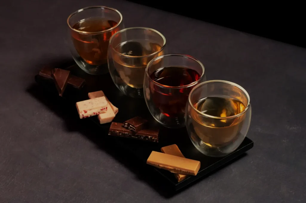 Four glass cups with different teas are lined up on a dark tray, each paired with a distinct piece of chocolate, creating a gourmet tasting setup.
