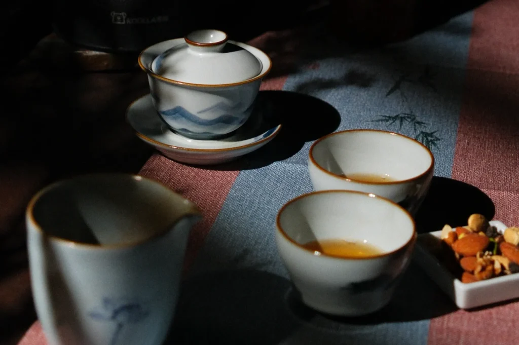 Ceramic tea set in dappled sunlight on a blue and red tablecloth. Two filled cups, a pot, and a dish with nuts create a calm, rustic ambiance.