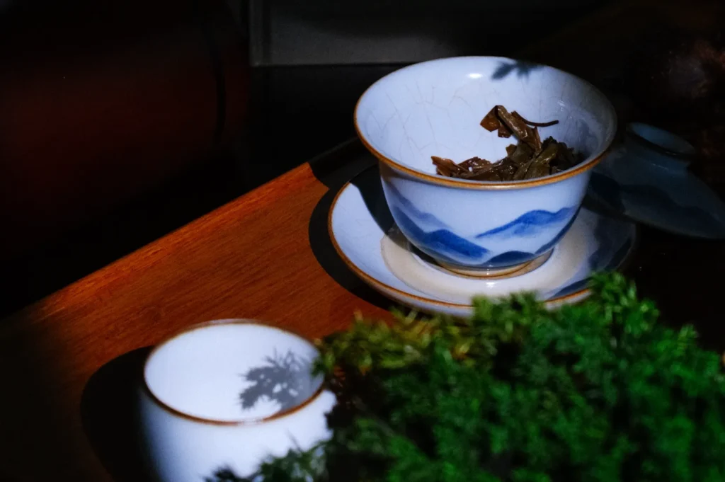 A bowl with tea leaves sits on a wooden surface, partially shadowed, alongside a small tea cup and some greenery.
