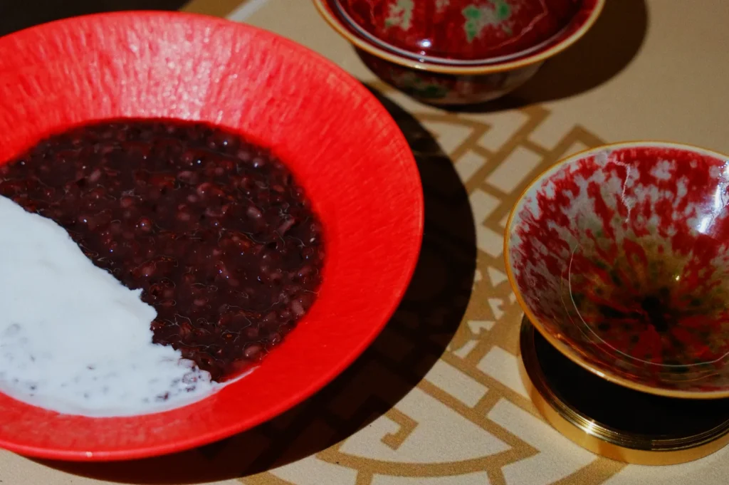 A vibrant red bowl holds a dish of dark red bean porridge with a splash of white creamy topping. Two decorative bowls are in the background on a patterned surface.