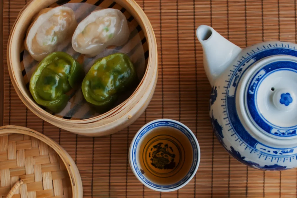 A bamboo steamer with two green and two white dumplings sits on a mat beside a blue and white porcelain teapot and a teacup filled with tea.