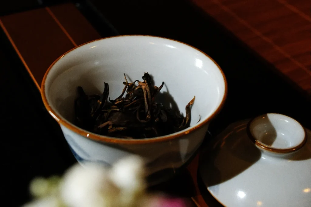 A small white tea cup with dried tea leaves inside while the lid rests on the side, partially covered by a flower.