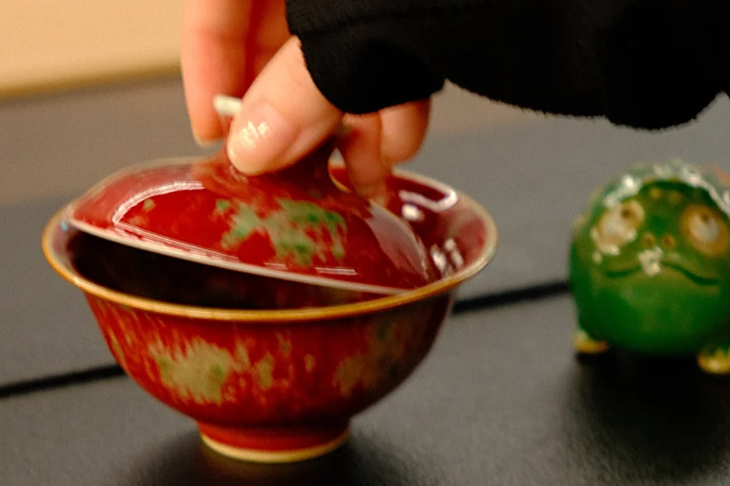 A hand wearing black long sleeves lifting the lid off a red tea cup, with a green frog figurine in the background.