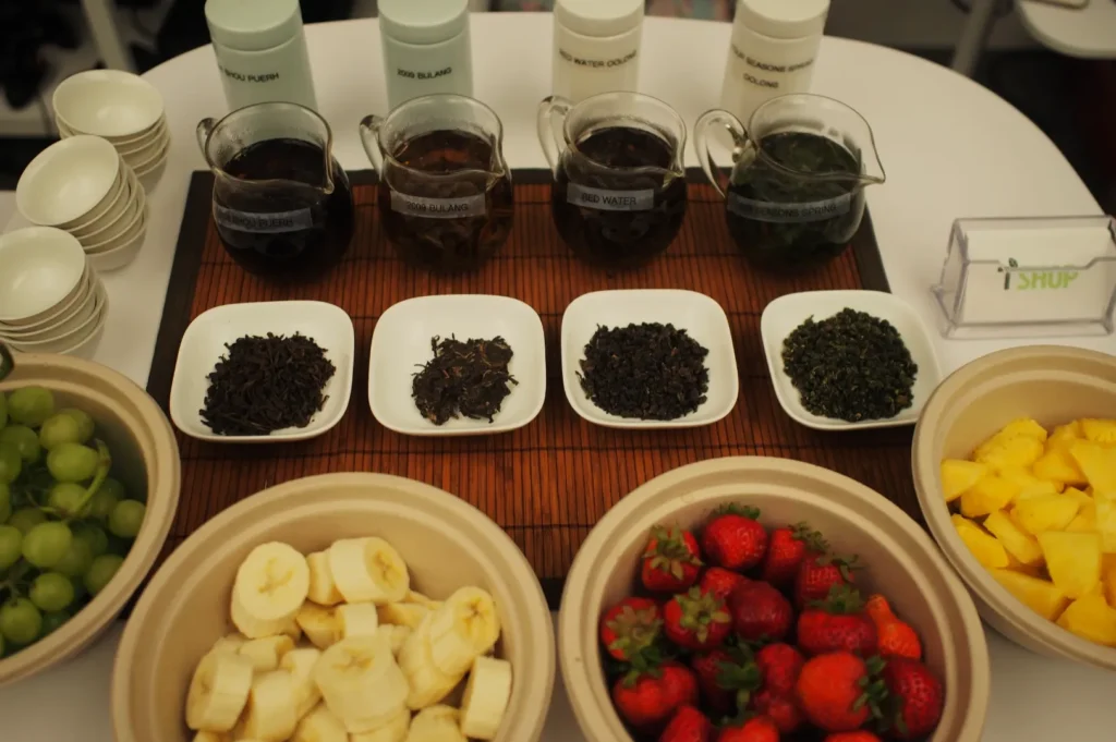 Assorted teas in glass pitchers with matching loose leaves on plates, surrounded by bowls of bananas, grapes, strawberries, and pineapple on a table.