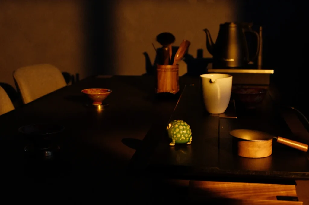 A dimly lit kitchen scene featuring a wooden table with various utensils, a ceramic bowl, a small green frog figurine, and a metal teapot in the background.