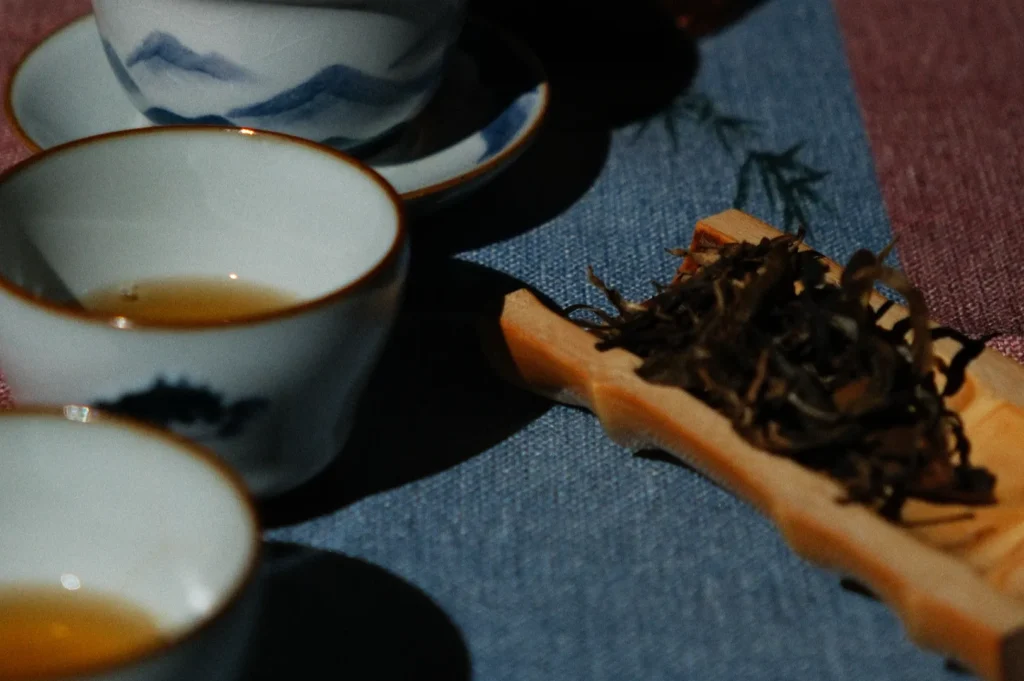 Three small white teacups with blue rims sit on a textured blue tablecloth, filled with light brown tea. Next to them, a bamboo tray holds dried tea leaves, adding to the tea setting's presentation.