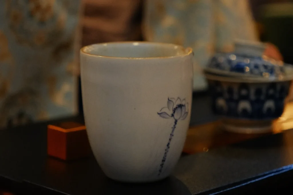 A ceramic cup with a delicate blue floral design sits on a table. In the background, there is a blue and white patterned dish, creating a serene atmosphere.