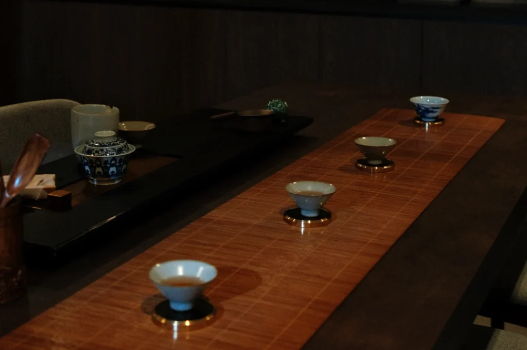 A dimly lit table set for traditional Japanese tea, with a bamboo mat and four intricate teacups. The atmosphere is serene and contemplative.