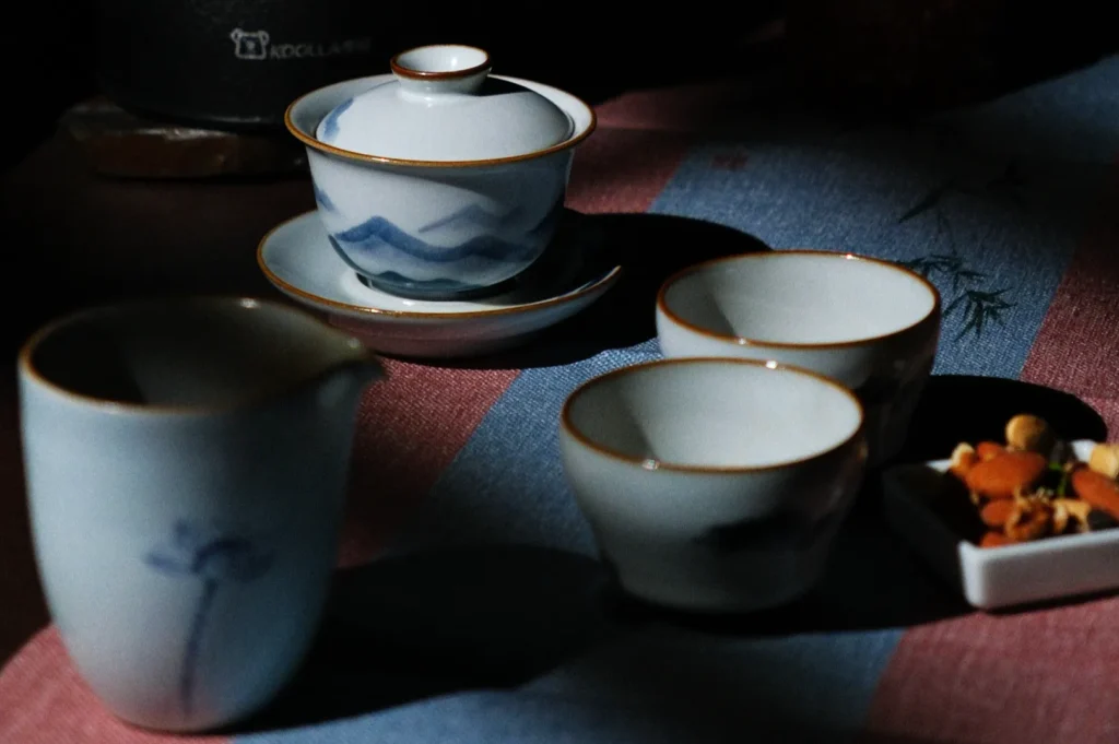 A still life image of elegant ceramic teacups, a gaiwan, and a pitcher on a patterned cloth, accented with nuts. The scene is softly lit, creating a calming and traditional atmosphere.