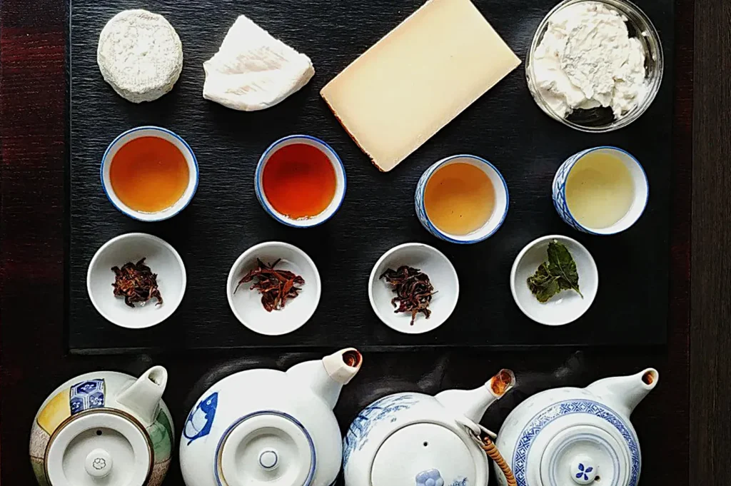 Assorted cheeses and loose leaf teas arranged on a dark board with three teapots below. Three cups of tea, each varying in color, sit alongside. Elegant and inviting.